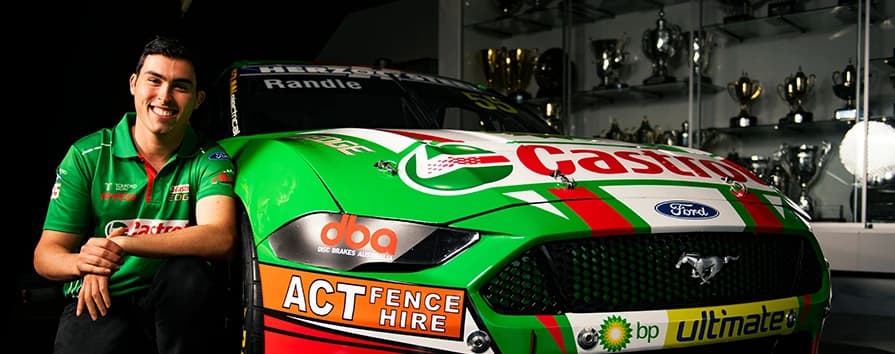 Professional motorsport driver with DBA-sponsored Supercars Championship vehicle showcasing racing partnership and performance braking technology Smiling race car driver in green team shirt standing next to DBA-sponsored green Ford Mustang racing car with multiple sponsor logos displayed in trophy room